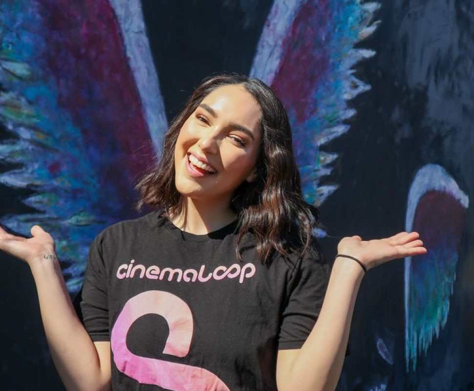 Cinemaloop Social media personality Female,, brown hair, white, smiling, hands fan up and back standing next to an angel wing graffiti in LA with a Cinemaloop T-shirt on